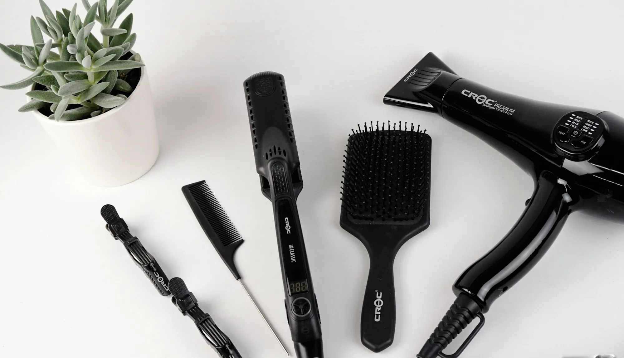 A black hairdryer, flat iron, comb and brush laid out on a white surface